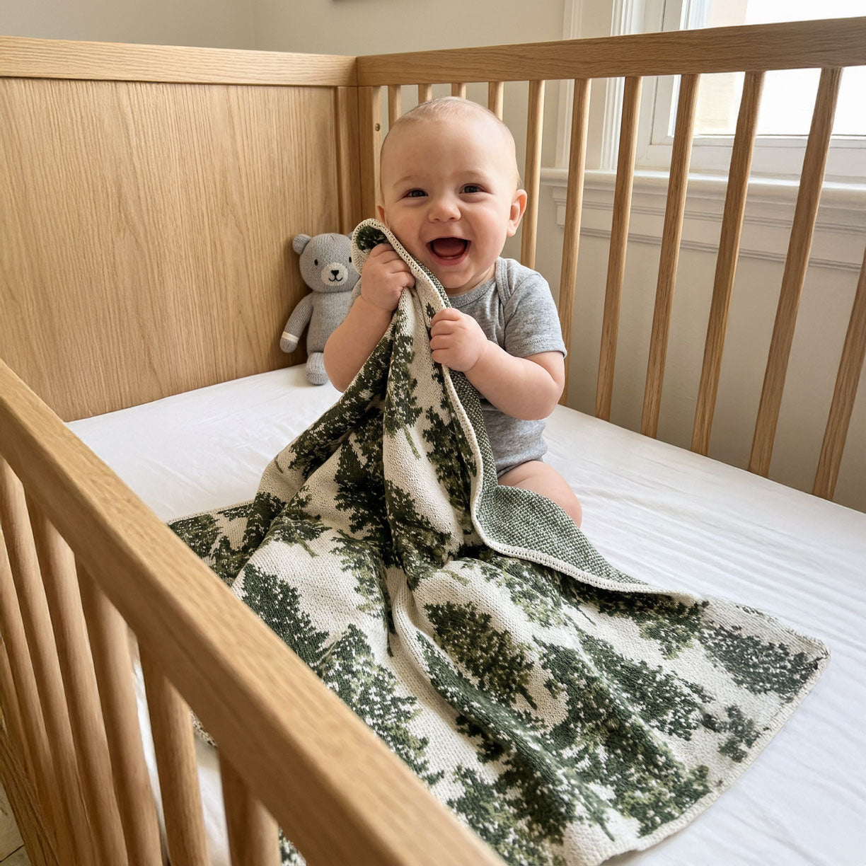Baby sitting in a crib with a green and white patterned blanket and a teddy bear.