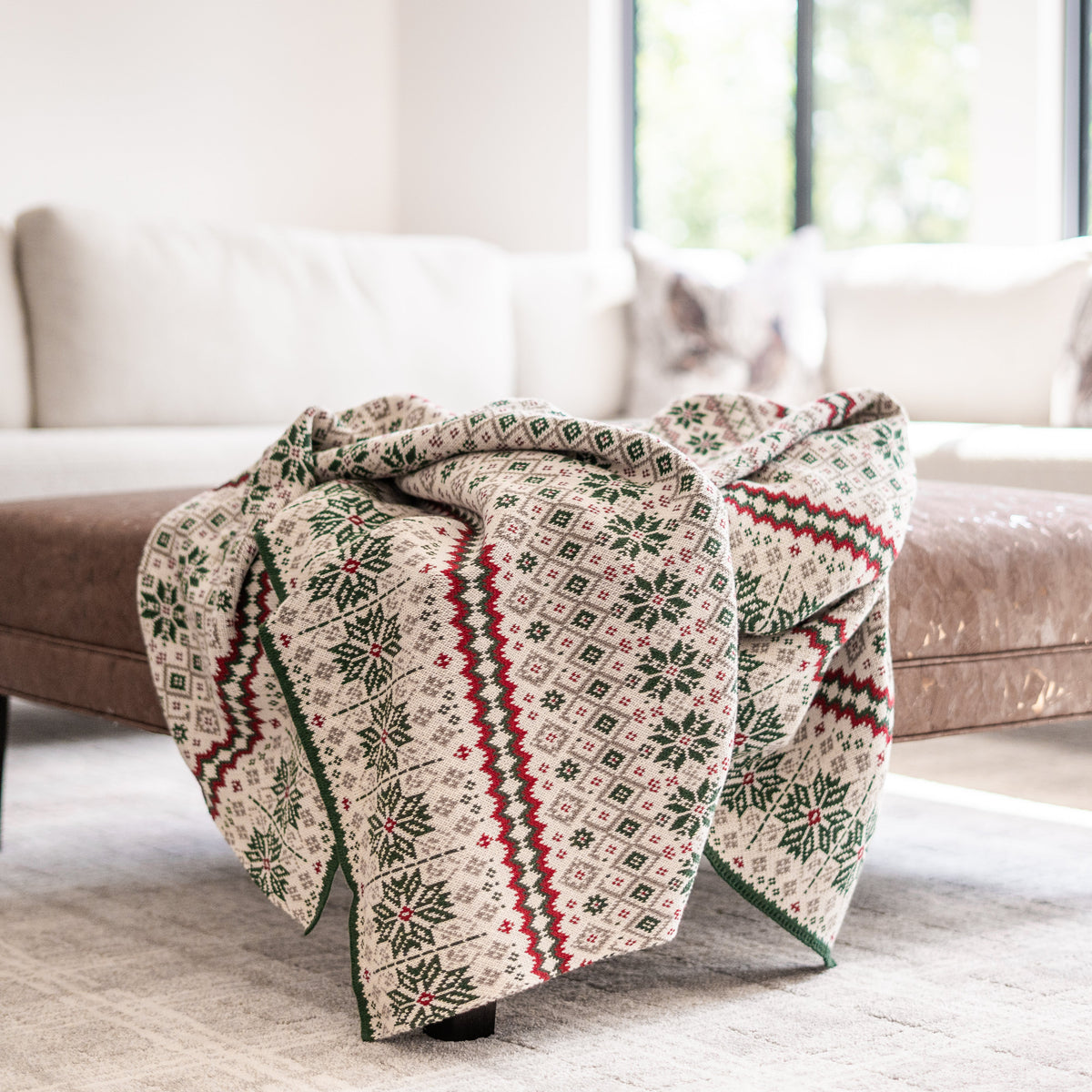 A cozy knit throw blanket with a festive Nordic pattern in green, red, and cream is draped over a brown leather ottoman in a bright, modern living room.
