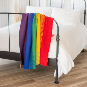 Seek & Swoon Rainbow Pride Throw blanket on a black iron bed with white linens